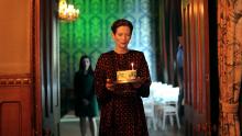 A woman stands in the doorway of a grand home, holding a birthday cake. Another woman stands behind her, out of focus.
