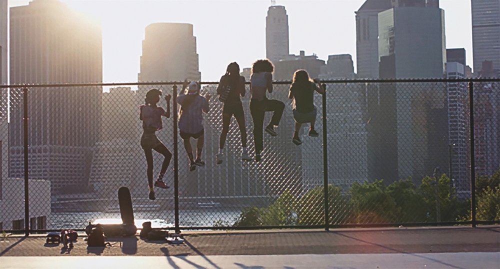 Image is from the film 'Skate Kitchen' (2018). Five young girls climb a chainlink fence. They are silhouetted against the New York City skyline.